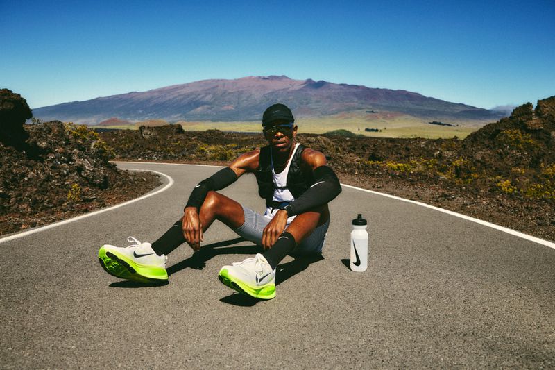 An African American male runner sits on a road with running shoes, a water bottle, and a mountain in the background under a blue sky.