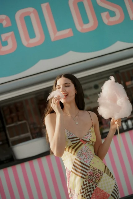 Young woman in summer dress enjoying pink cotton candy by a vintage food truck with the text 'COLOSSO' on its side.