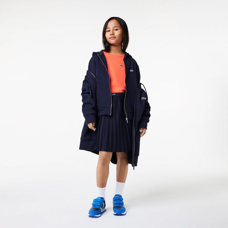 Child model wears Lacoste outfit with a navy jacket, pleated skirt, orange t-shirt, and blue sneakers against a white backdrop.