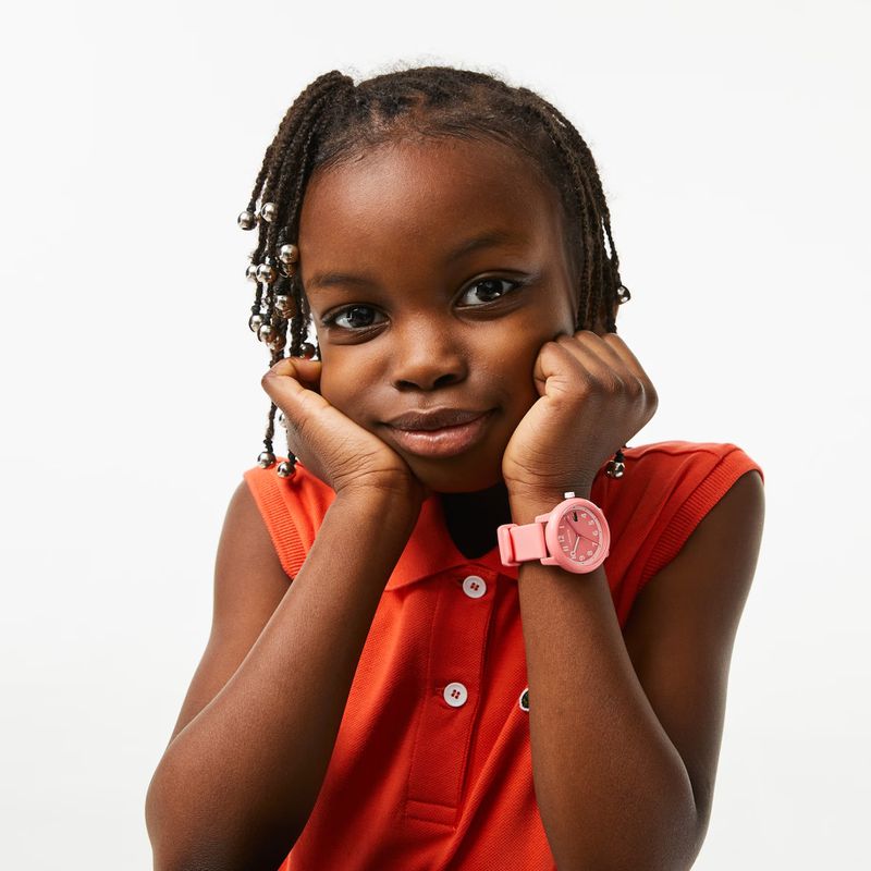 Portrait of a young Black girl in a red polo shirt, resting her chin on her hands and wearing a pink wristwatch against a white backdrop.