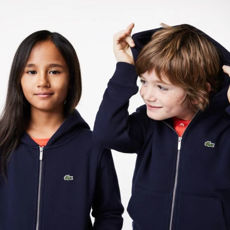 Two children, a girl and a boy, are posing indoors, wearing matching navy zip-up hooded sweaters with the Lacoste logo on the chest.