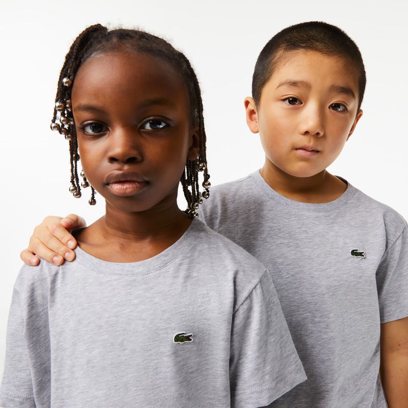 Two children, a girl and a boy, in gray t-shirts featuring the Lacoste logo, pose in a studio setting with a clean white background.