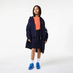 Young child model wearing a navy hoodie over an orange top and pleated skirt with blue sneakers against a white background