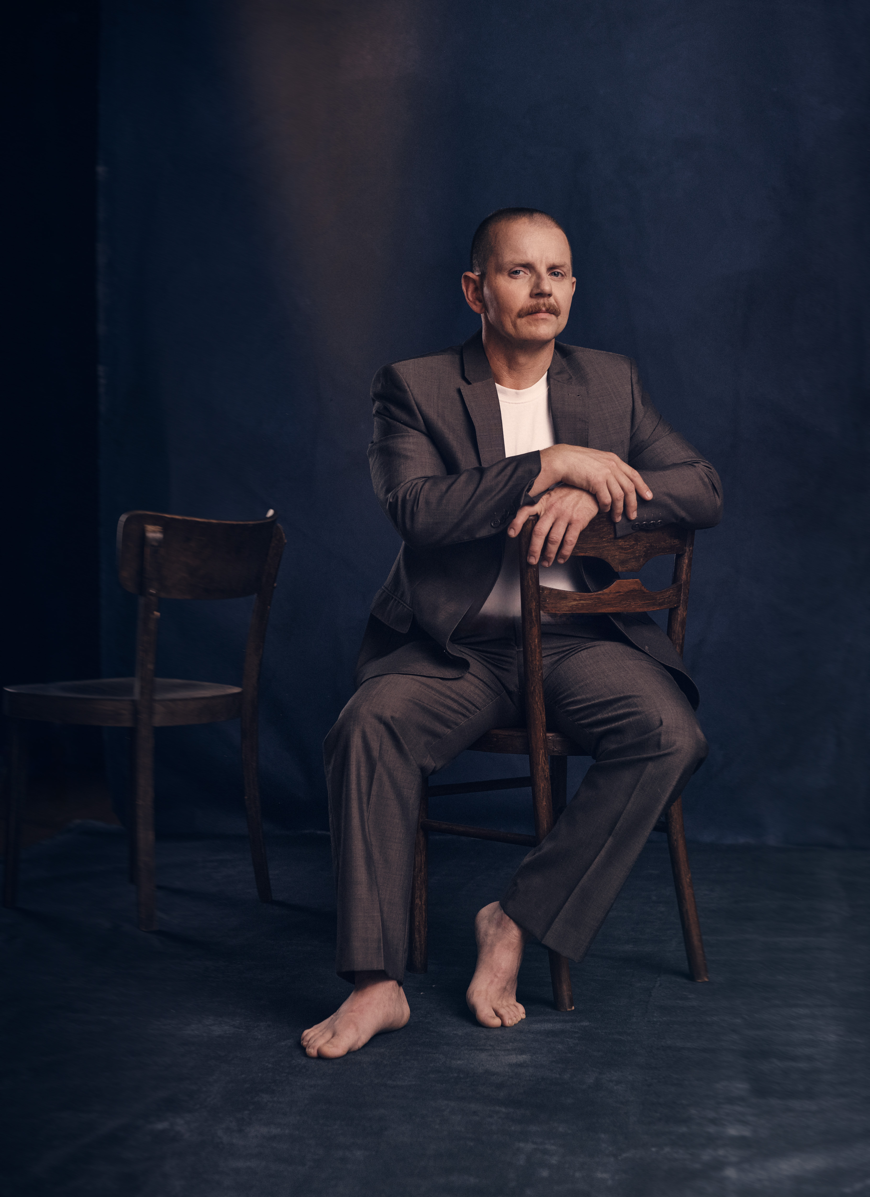 A barefoot man with a mustache in a grey suit sits backwards on a wooden chair against a dark moody studio backdrop.