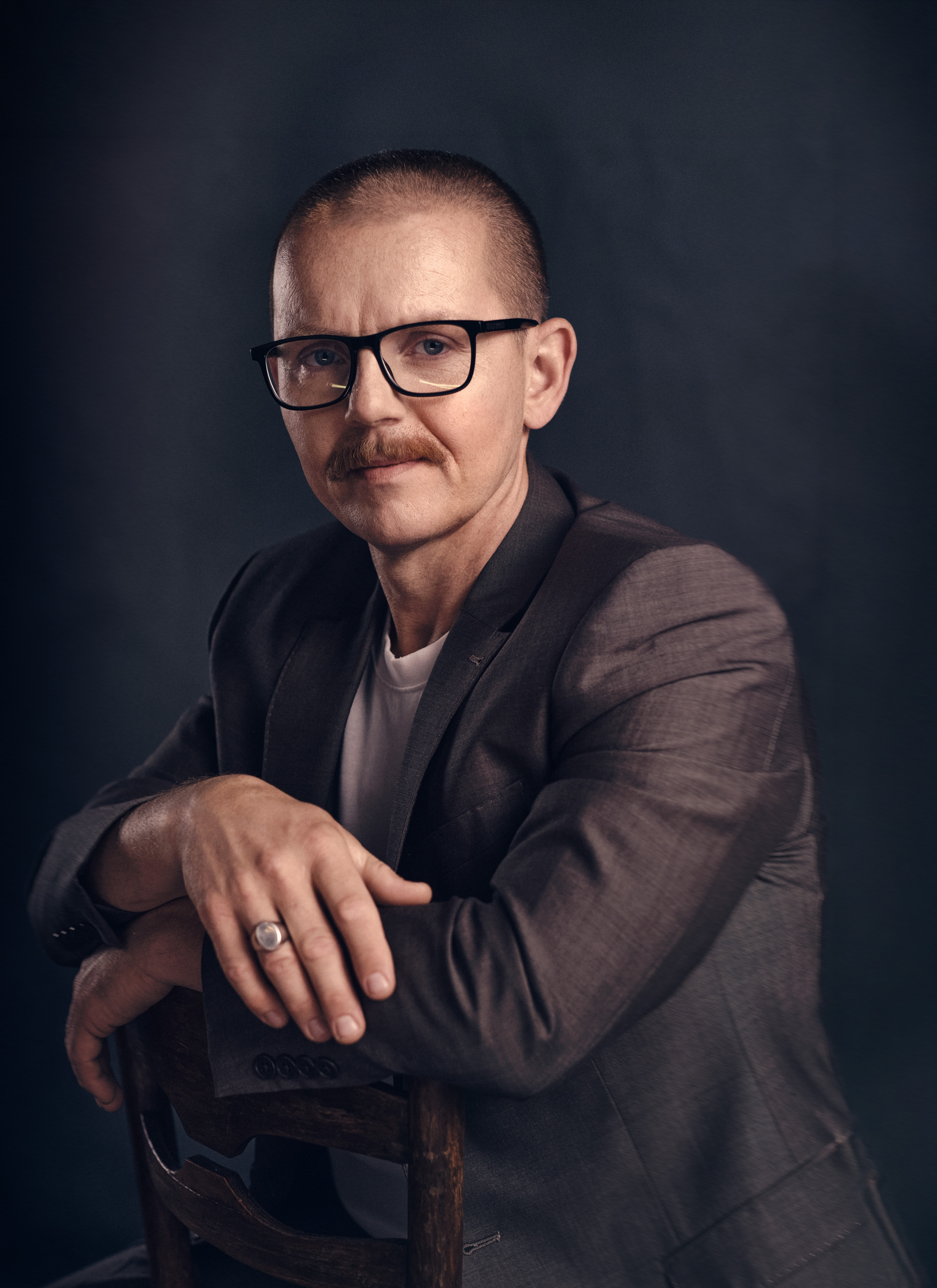 Portrait of a middle-aged man with glasses, mustache, and buzzcut, wearing a gray suit and seated on a wooden chair in studio lighting.