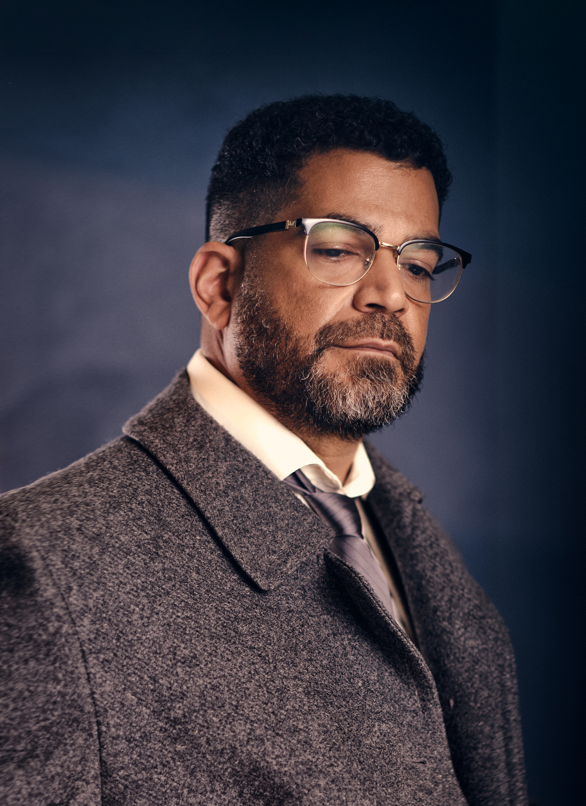 Portrait of a distinguished man with a beard wearing glasses, a gray coat, and a tie, looking down with a serious expression in a studio setting.