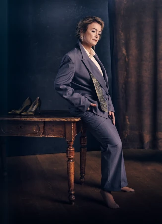 Mature woman poses in a grey suit, seated on an old wooden desk with high heels, against a muted backdrop, creating a sophisticated portrait.