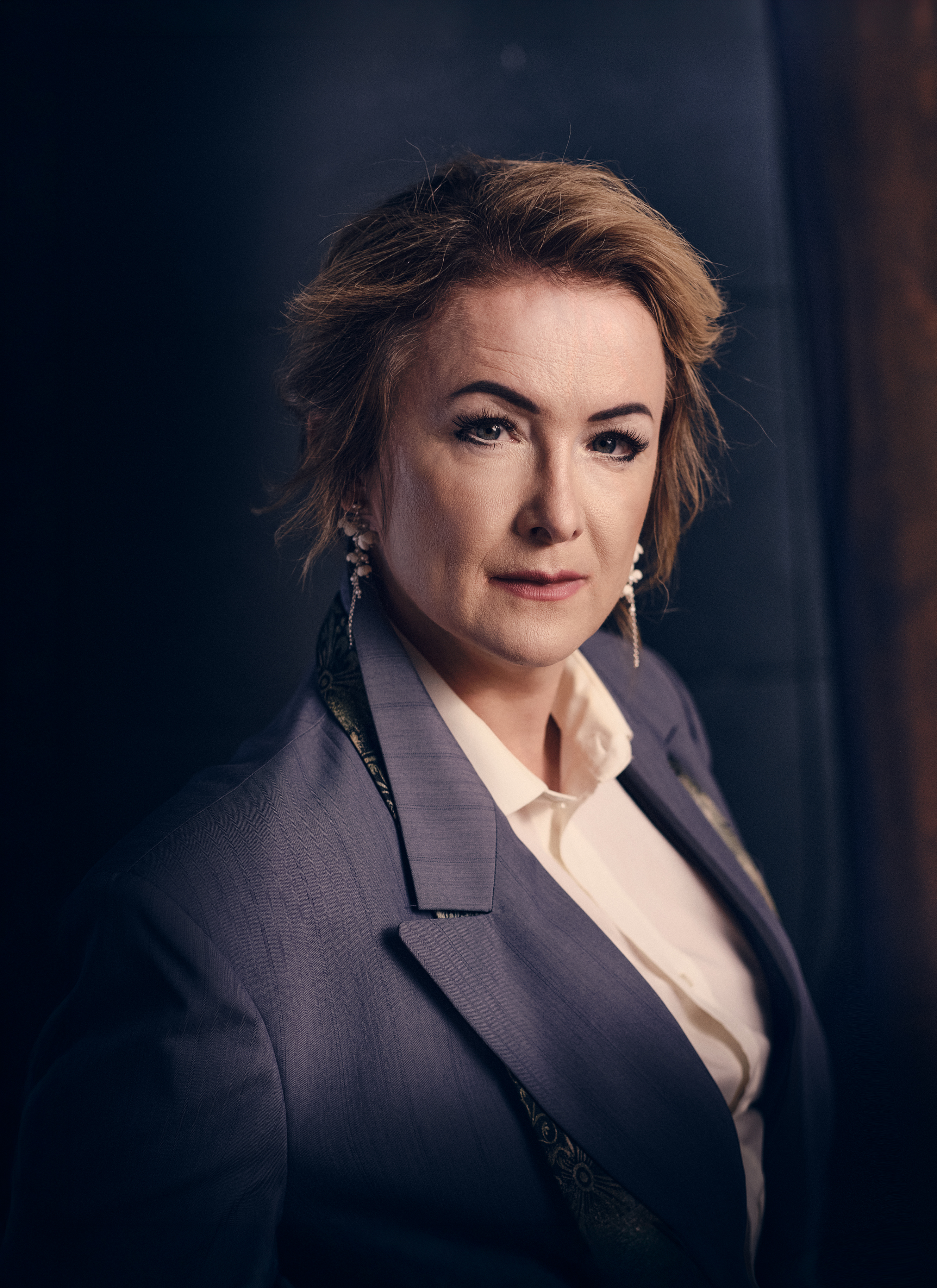 Mature woman in a grey blazer and white shirt stands in a portrait. She has short hair and dangle earrings, shot in a studio.