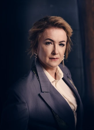 Mature woman in a grey blazer and white shirt stands in a portrait. She has short hair and dangle earrings, shot in a studio.
