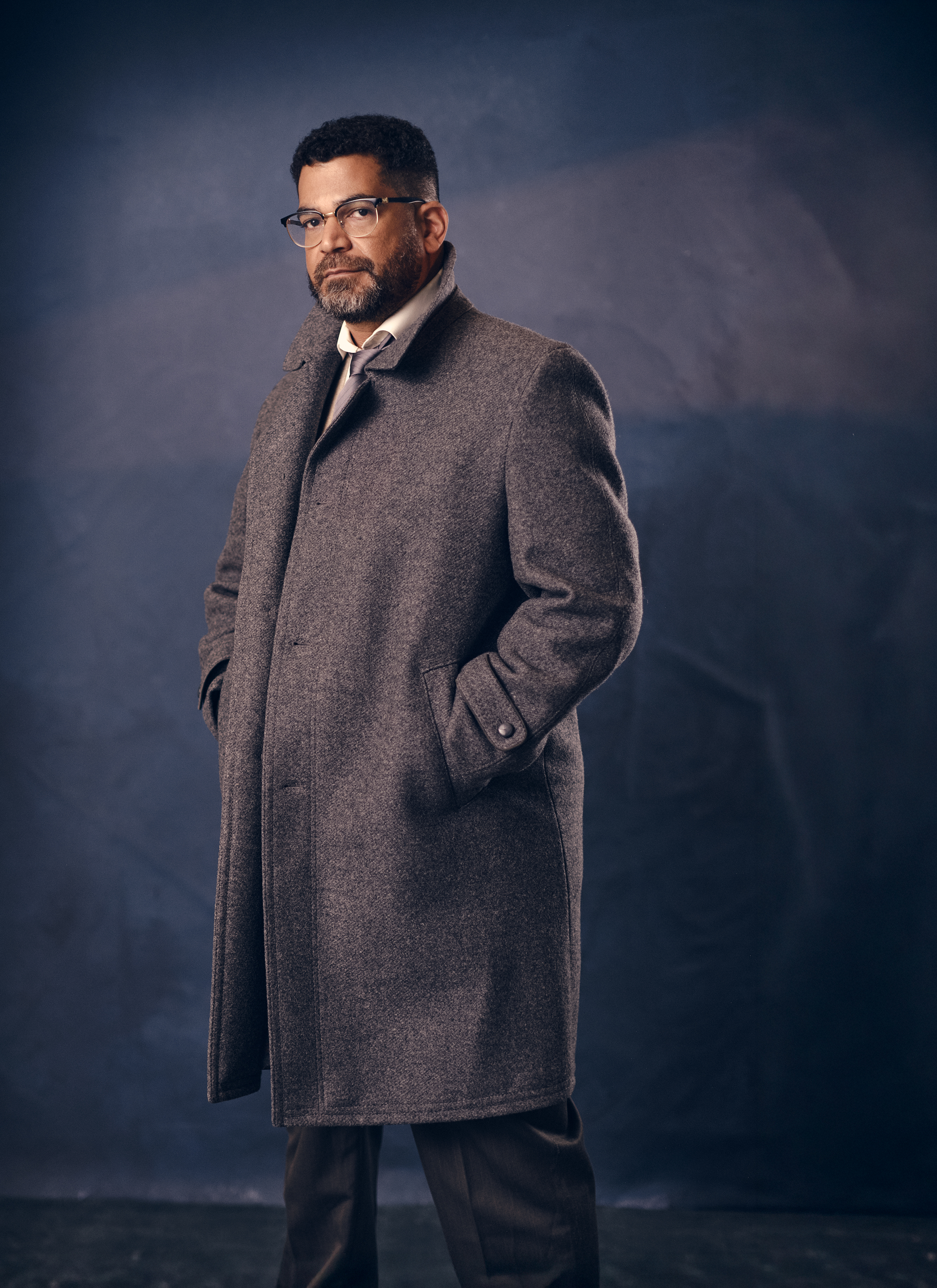 Portrait of an adult male with glasses wearing a grey coat and trousers against a dark blue, mottled background in a studio setting.