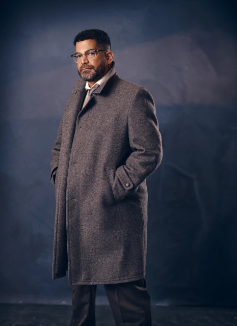 Portrait of an adult male with glasses wearing a grey coat and trousers against a dark blue, mottled background in a studio setting.