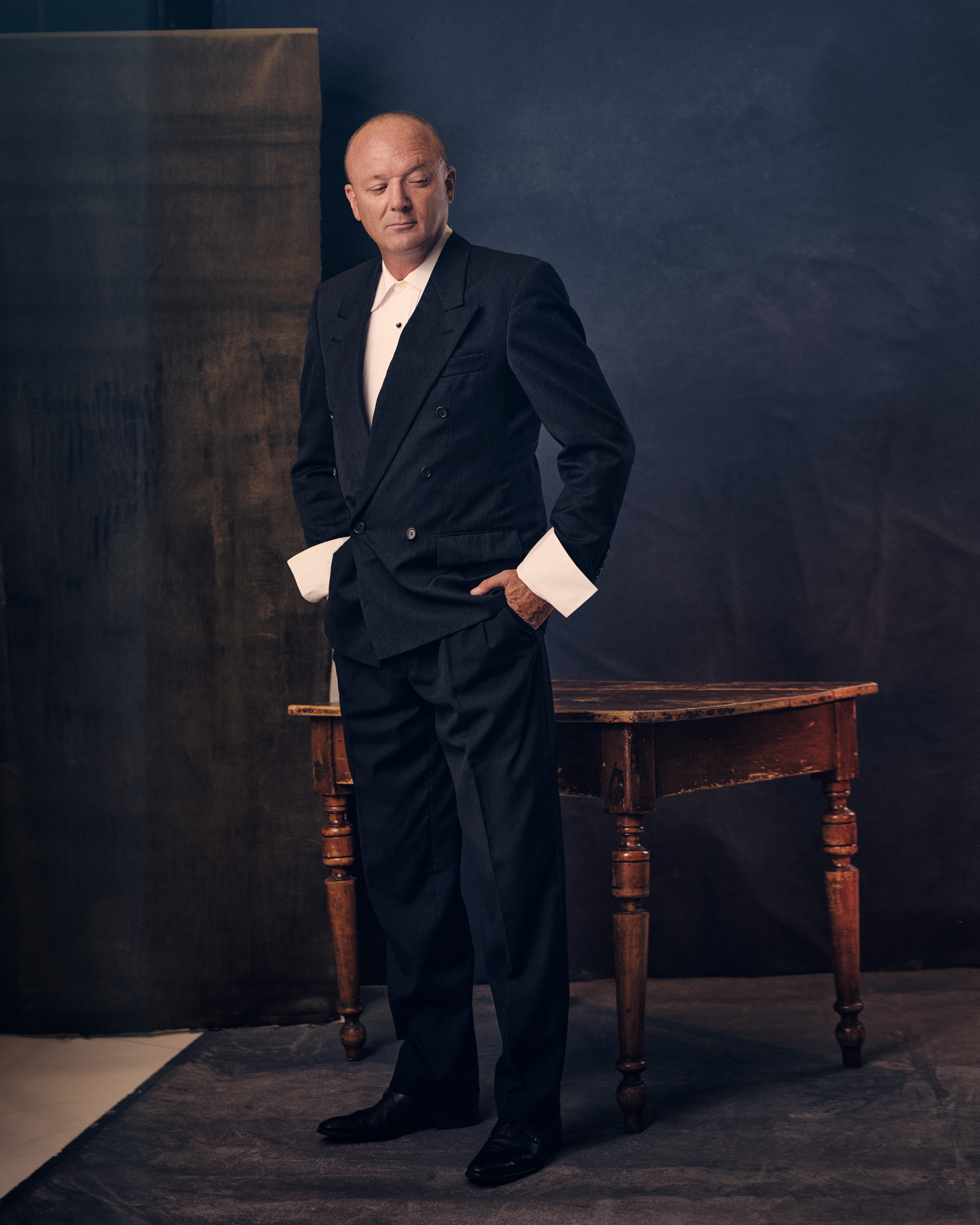 A mature man in a dark suit with wide lapels poses near a wooden table against a dark background in a studio portrait.