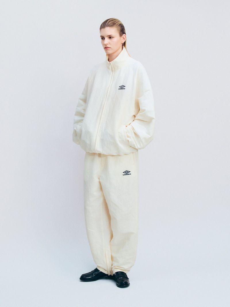 Woman in a cream-colored Umbro tracksuit stands in a white studio, captured in a full shot. Athletic wear fashion against a clean backdrop.