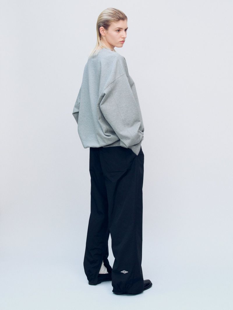 Blonde woman in gray sweatshirt and black pants stands in a neutral studio, looking over her shoulder toward the light.