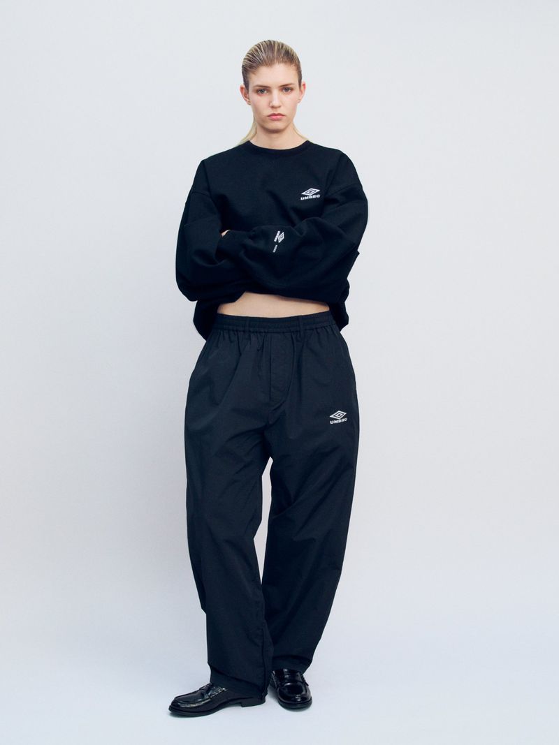 Full-length studio shot of a female fashion model in a black long-sleeve sweatshirt, wide black pants, and black patent-leather shoes.