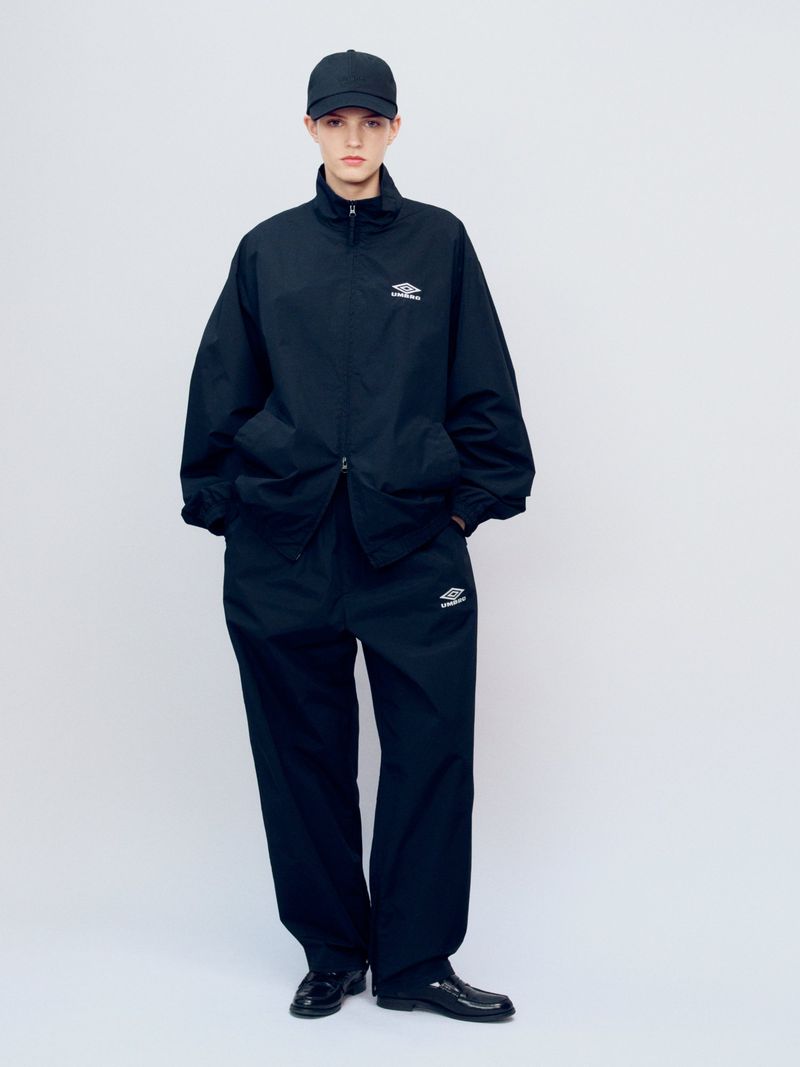 Full-length shot of a person wearing a black tracksuit with logo, baseball cap, and patent leather shoes against a plain studio backdrop.