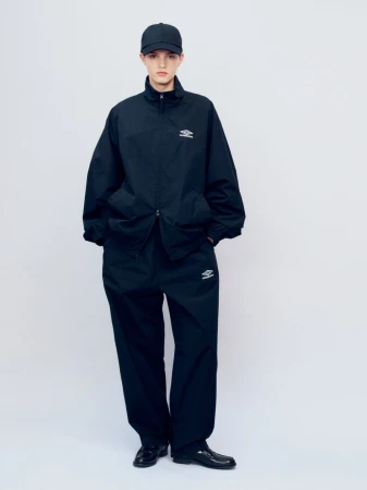 Full-length shot of a person wearing a black tracksuit with logo, baseball cap, and patent leather shoes against a plain studio backdrop.