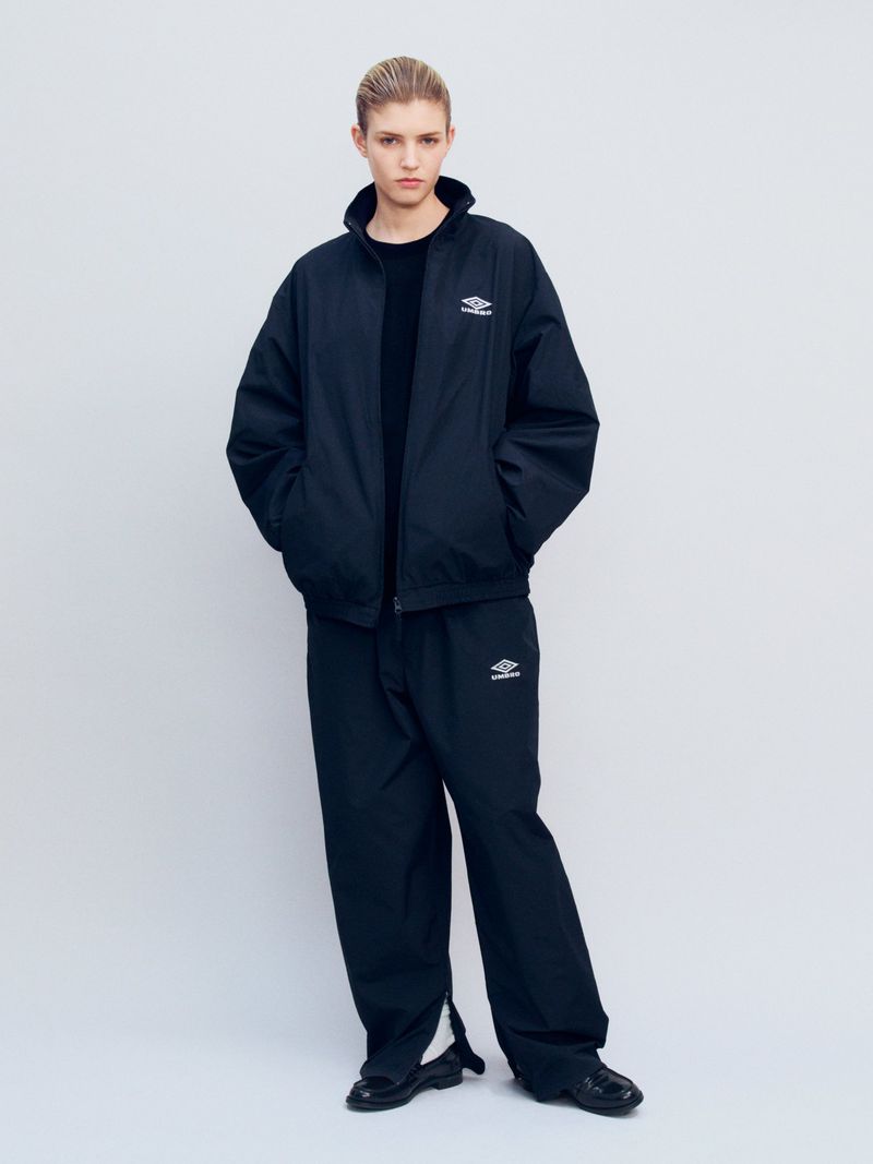 An adult female fashion model poses in a black Umbro windbreaker jacket and black sweatpants against a solid white backdrop.