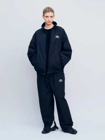 An adult female fashion model poses in a black Umbro windbreaker jacket and black sweatpants against a solid white backdrop.