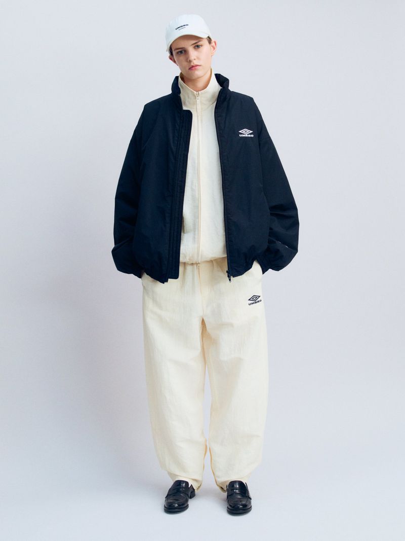 Model wearing a black zip jacket over a cream tracksuit and white cap, standing against a white studio backdrop.