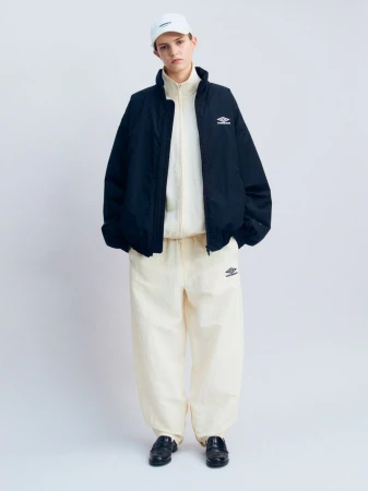 Model wearing a black zip jacket over a cream tracksuit and white cap, standing against a white studio backdrop.
