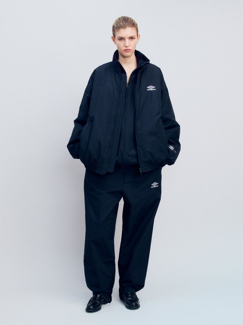 A woman stands in a light grey studio wearing a navy Umbro tracksuit and black shoes, shot in full length with soft lighting.