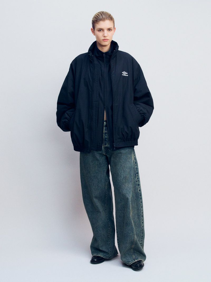 A woman poses in a dark blue Umbro jacket and wide-leg jeans against a pale backdrop, showcasing casual fashion in a studio setting.