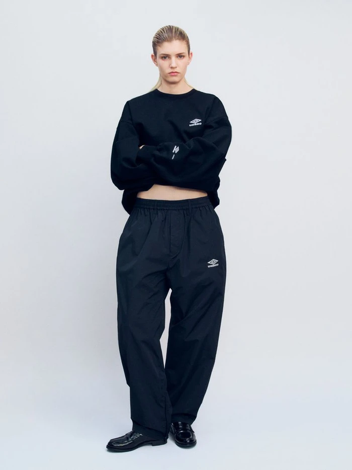 Model Camille Chifflot standing in an oversized black Umbro sweatshirt and matching black track pants against a white background.