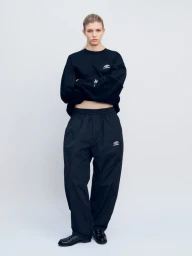 Model Camille Chifflot standing in an oversized black Umbro sweatshirt and matching black track pants against a white background.