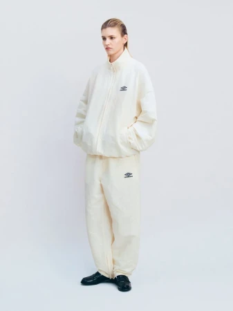A model standing in a three-quarter pose wearing an oversized cream ssstein x Umbro tracksuit and black loafers against a white backdrop.