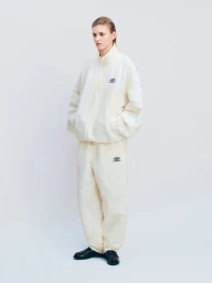 A model standing in a three-quarter pose wearing an oversized cream ssstein x Umbro tracksuit and black loafers against a white backdrop.