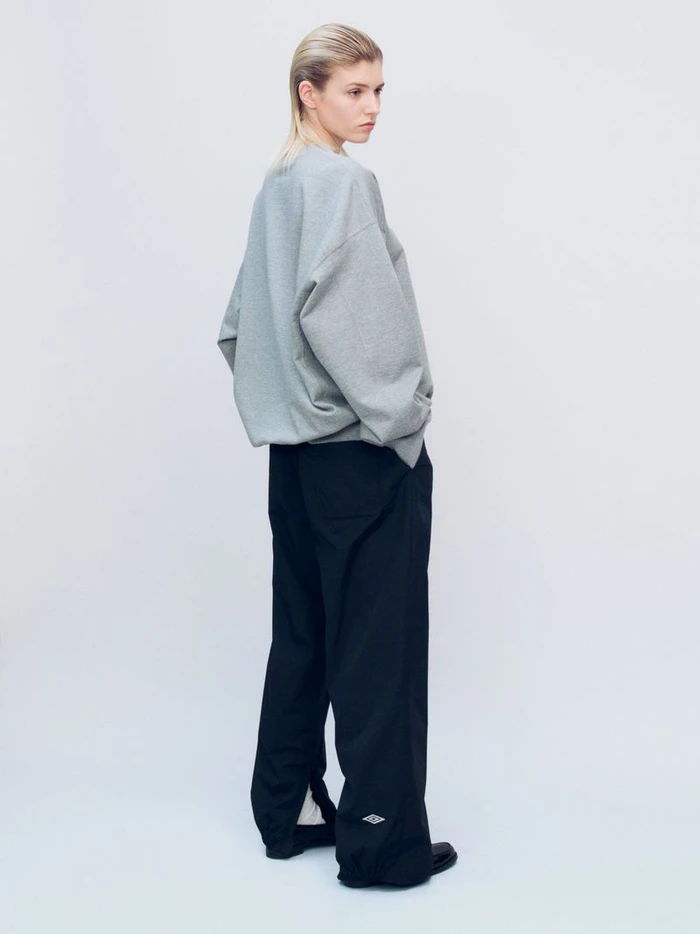 Model wearing an oversized light grey sweatshirt and loose black track pants viewed from behind against a neutral studio backdrop.