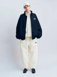 Model wearing a black zip jacket over a cream tracksuit and white cap, standing against a white studio backdrop.