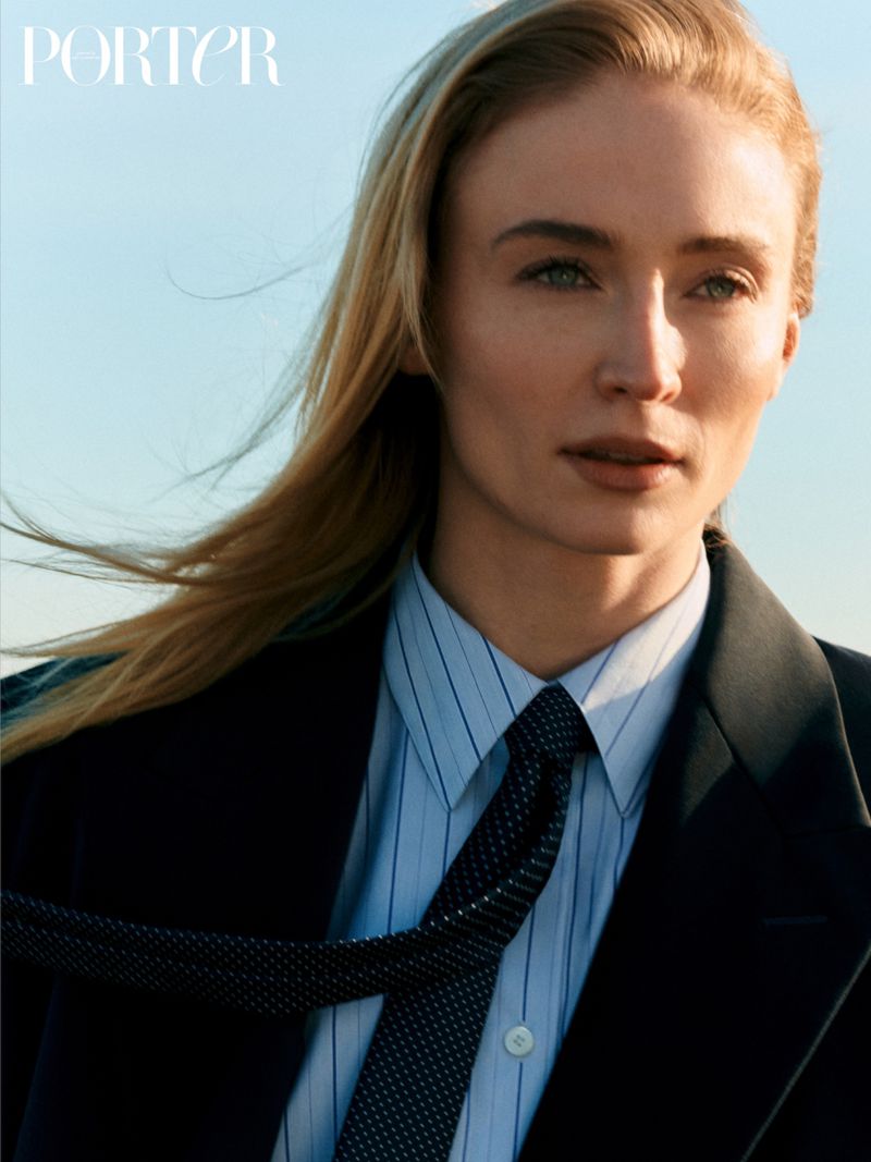 Portrait of a blonde woman wearing a blue and white striped shirt, black jacket, and dark tie against a light blue background.