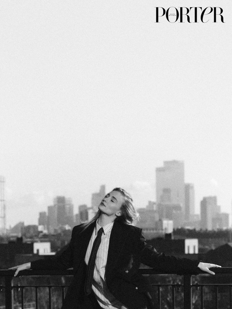 Monochrome fashion portrait of a blonde woman in a suit and tie outdoors with a city skyline backdrop on a rooftop.