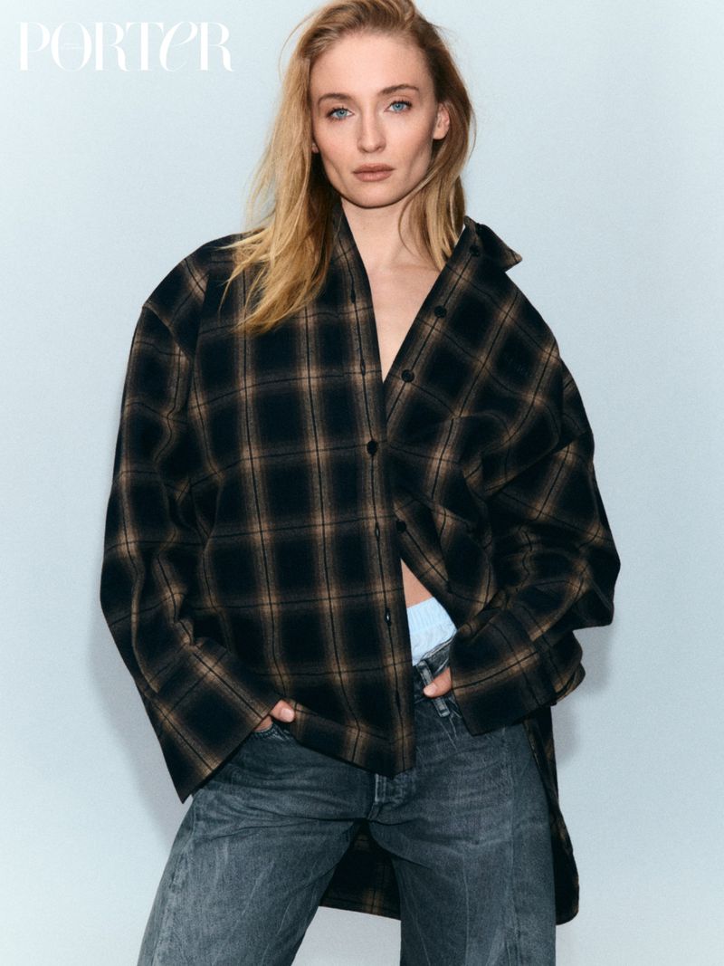 Fashion editorial portrait of a blonde woman wearing a plaid jacket and jeans, standing in front of a white studio backdrop with natural lighting.