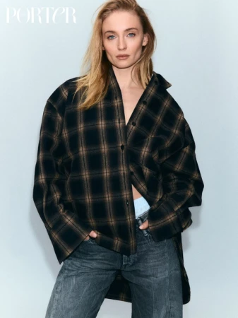 Fashion editorial portrait of a blonde woman wearing a plaid jacket and jeans, standing in front of a white studio backdrop with natural lighting.