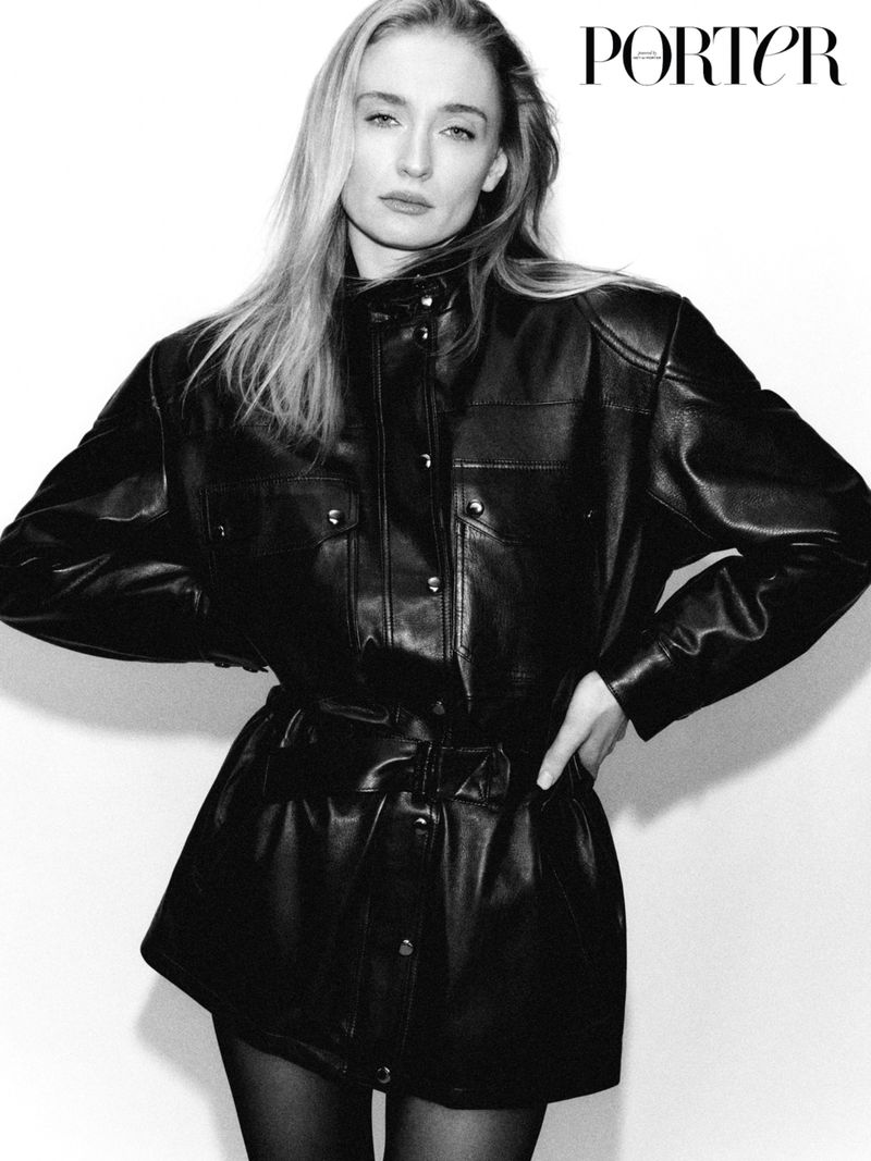 Black and white portrait of a blonde woman wearing a black leather button-down dress with a belt against a white backdrop.