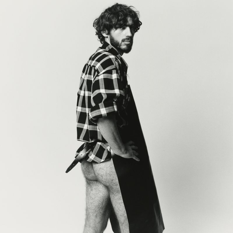 Portrait of a bearded adult male posing in a studio wearing a plaid shirt and black apron covering his lower body against a white backdrop.