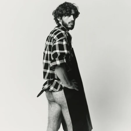Portrait of a bearded adult male posing in a studio wearing a plaid shirt and black apron covering his lower body against a white backdrop.