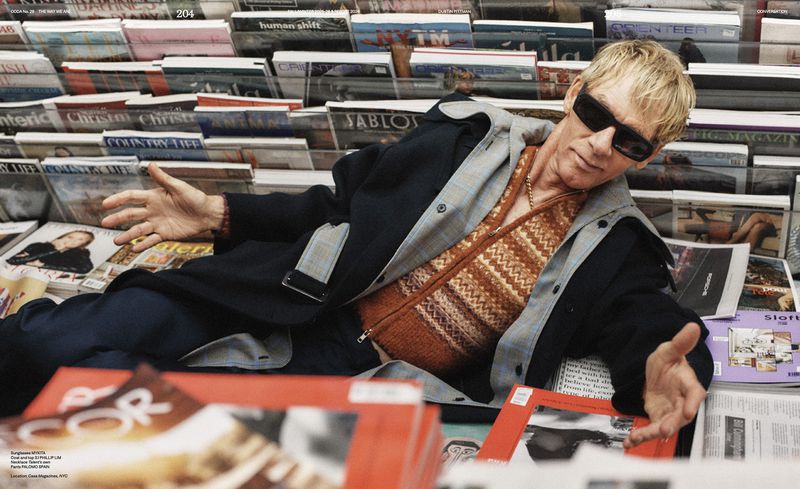 Adult male lying down, surrounded by stacks of magazines, wearing a patterned vest, sunglasses, and jacket in an indoor setting.