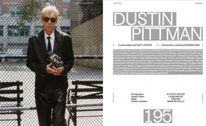 Dustin Pittman wearing a black leather suit and dark sunglasses while holding a vintage camera against a chain-link fence on a magazine page.