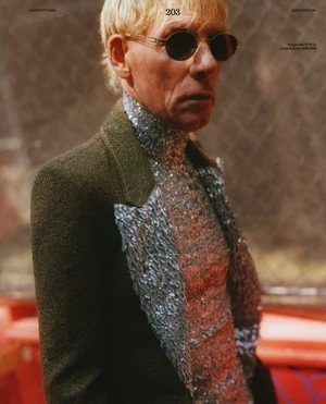 Dustin Pittman wearing dark sunglasses, a silver sequin turtleneck, and an olive green blazer in profile.