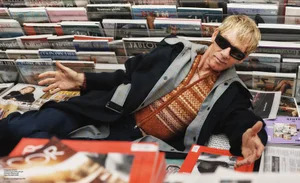 Man wearing sunglasses and layered winter clothing reclining amidst a display of magazines and books.
