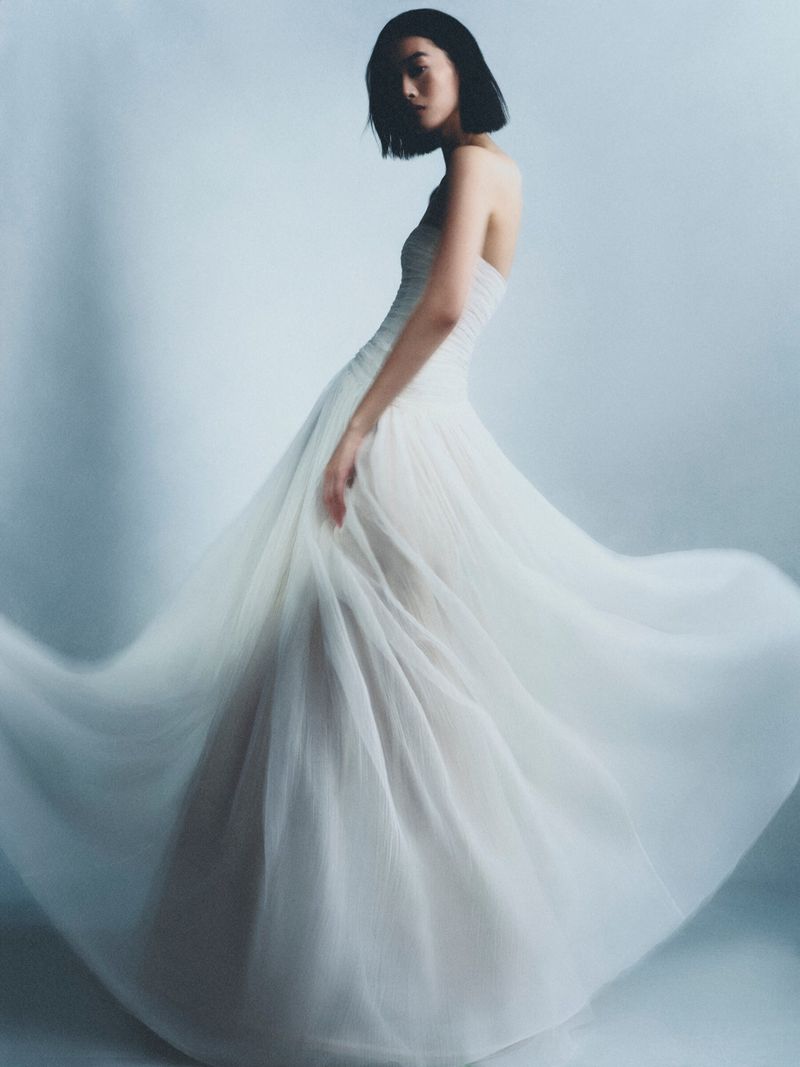 Elegant female fashion model in a flowing strapless white dress. The image is a medium shot against a soft, neutral background.