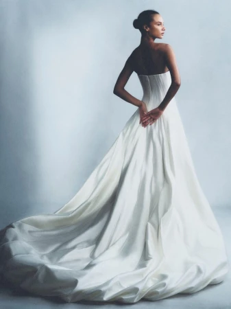 Elegant portrait of a slender adult female in an off-the-shoulder white wedding dress with hands clasped behind her back in a studio.