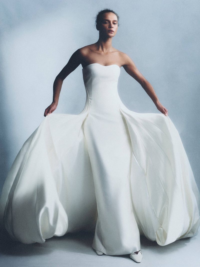 Adult female poses wearing a white strapless gown in a studio. The dress has a fitted bodice and large puff sleeves at the waist.