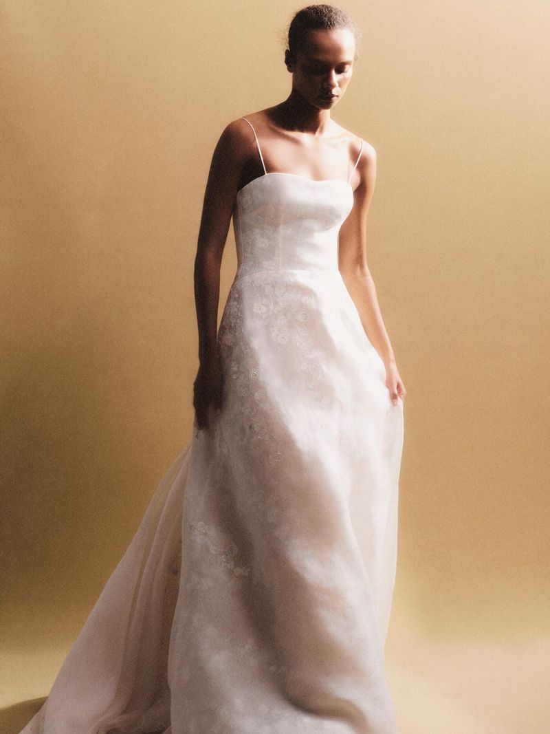 Elegant woman in a white gown with spaghetti straps and subtle floral patterns against a warm backdrop, creating a serene bridal aesthetic.