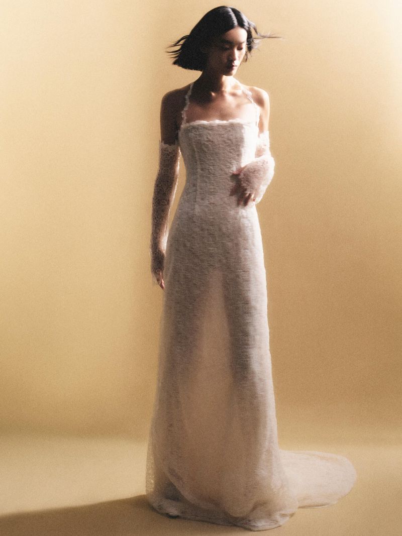 A woman with short black hair wears a floor-length white gown and sheer arm coverings against a warm, monochromatic backdrop in soft studio lighting.