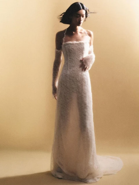 A woman with short black hair wears a floor-length white gown and sheer arm coverings against a warm, monochromatic backdrop in soft studio lighting.